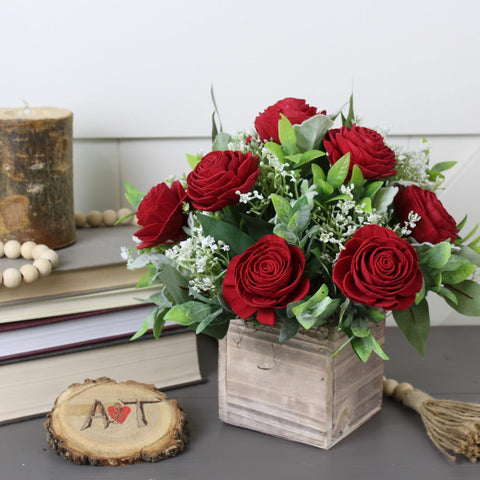 A dozen red sola wood roses sets on a table with some books, and a candle.