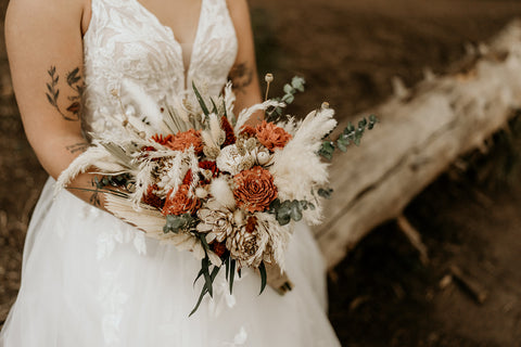A bride holds a boho style bridal bouquet made with ivory, rust, and terracotta sola wood flowers. 