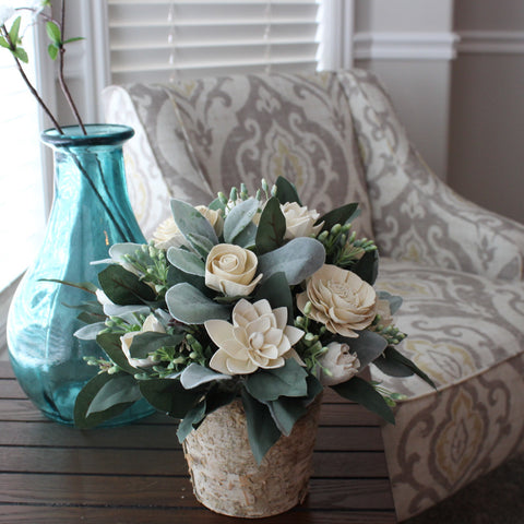 White sola wood flowers in a birch-wrapped tin sit on a table with a blue vase. 