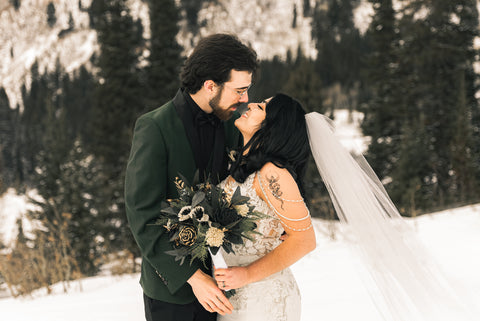 A bride and groom gaze at each other in a snowy, winter meadow. She is holding a sola wood flower bridal bouquet with black sola wood roses, white sola wood flowers, with gold accents.