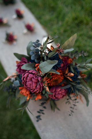 A maroon, rust, and navy sola wood flower bridal bouquet sets on a rock wall.
