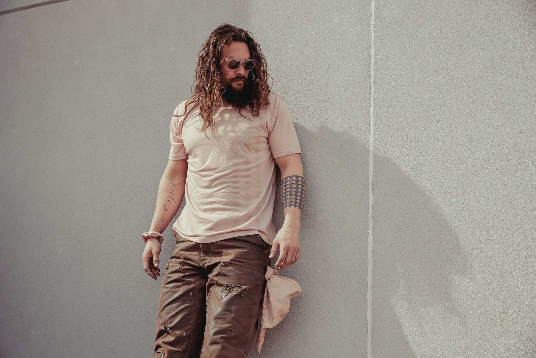 Jason Momoa shown wearing the so ill x on the roam dirty pink nokoa tee leaning up against a wall