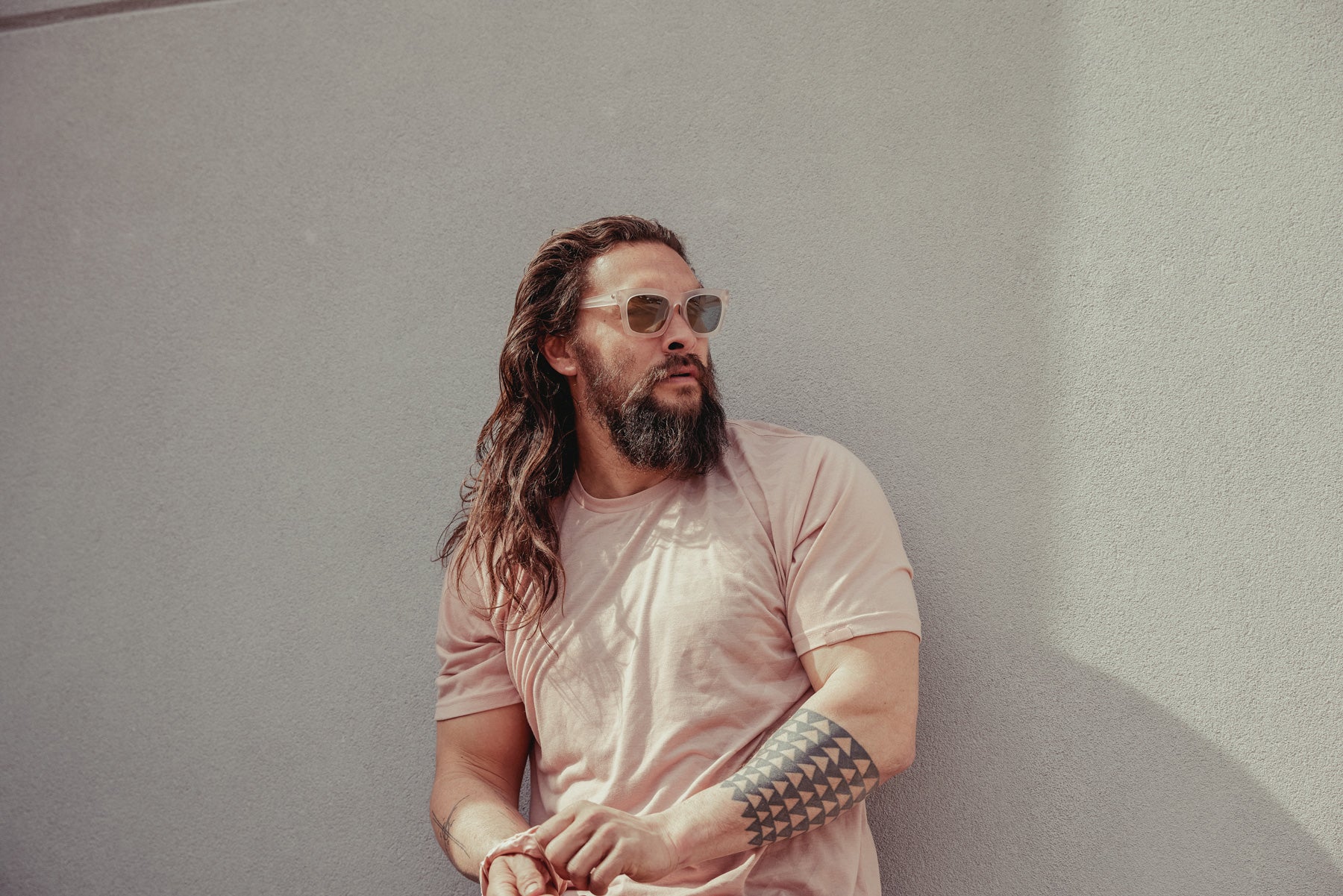 Jason Momoa shown wearing the so ill x on the roam dirty pink nokoa tee leaning up against a wall