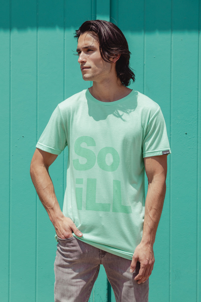 A man wears the seafoam colored stacked logo tee in front of a teal door