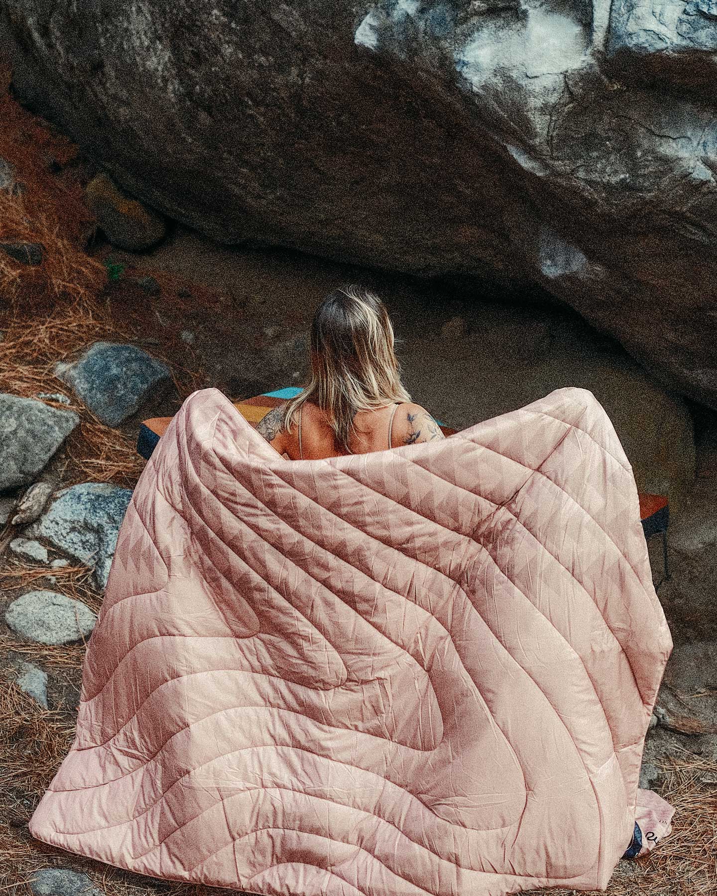 A woman sitting in front of a boulder, with the Dirty Pink Original Puffy Blanket from the Rumpl x So iLL x On The Roam, by Jason Momoa collaboration draped around her back.