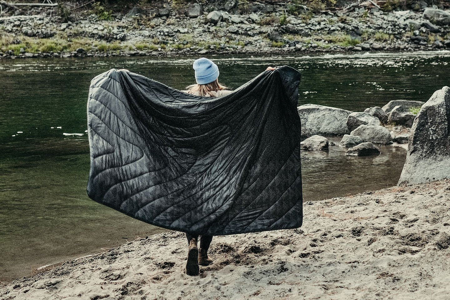 A woman walking down a beach while holding the Black Wolf Original Puffy Blanket from the Rumpl x So iLL x On The Roam, by Jason Momoa collaboration out behind her.