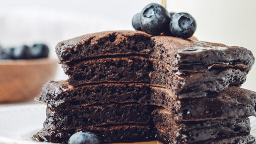 Syrup being poured onto a stack of chocolate buttermilk pancakes with blueberries