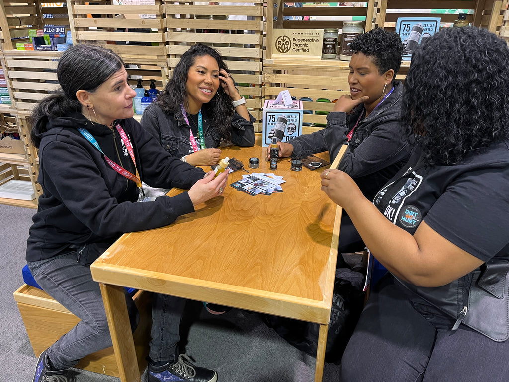 four women, a Dr. Bronner's employee and the staff of Dirt Don't Hurt talking to each other at a table at a Natural Products Expo.