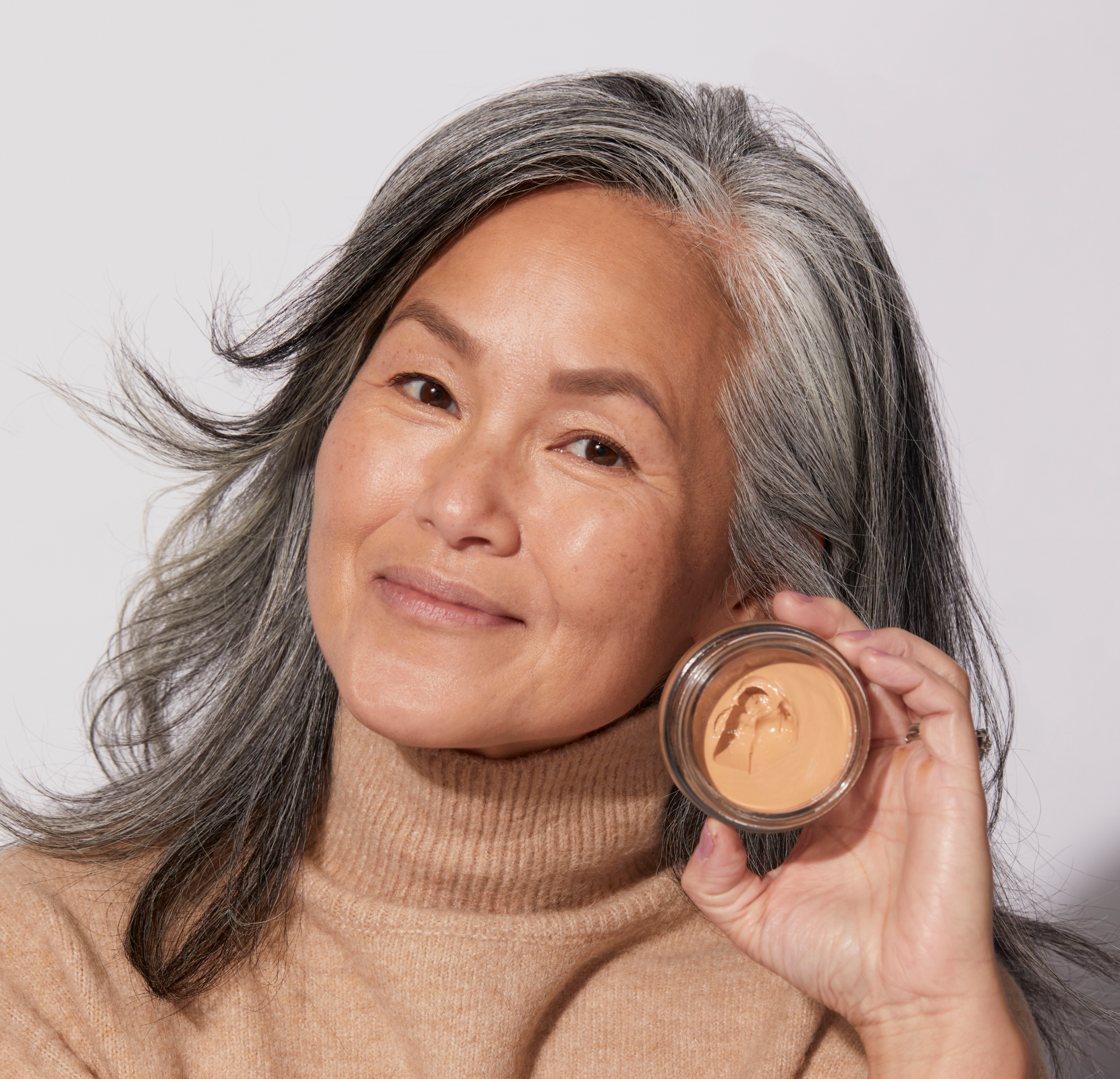 Woman with gray hair holding a container of skincare cream, smiling.