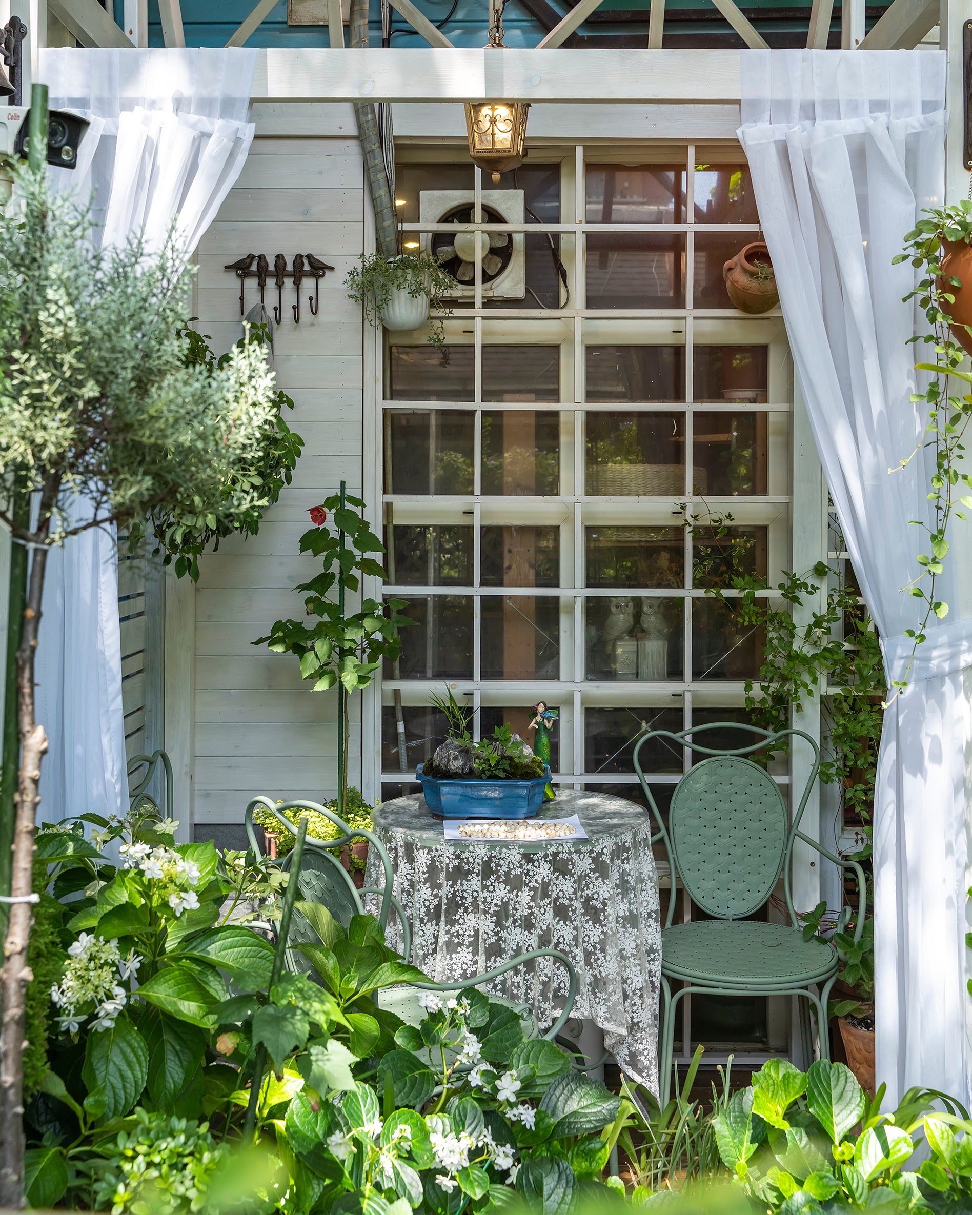 Looking at the back porch from outside. There is a little metal table and chair decorated nicely, surrounded by lush green plants.