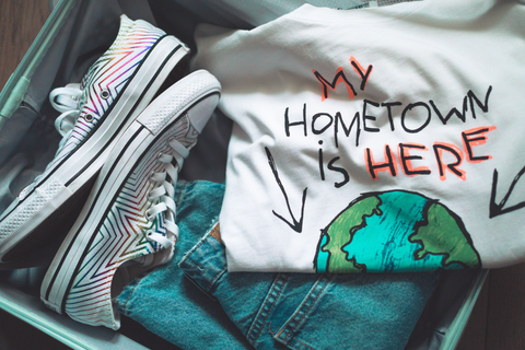 Suitcase filled with shoes, pants, and a t-shirt with a photo of a globe that reads “My Home is Here”. 