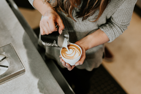 A photo of a woman barista pouring frothed milk and creating a pattern in a latte cup. 