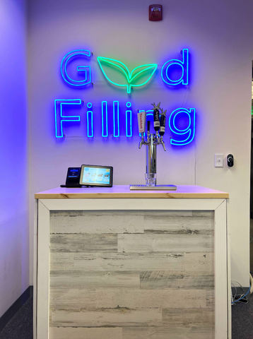 A photo of a self-service drink machine called a Hydration Station in front of a neon sign that reads “Good Filling”.