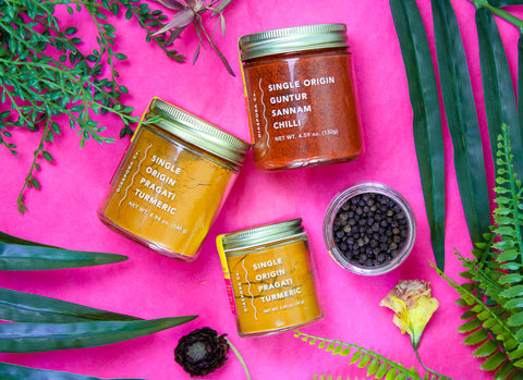 A smaller image of jars of spices made by Diaspora Co against a pink background. One small jar of turmeric, one small open jar of peppercorns, one large jar of turmeric, and one large jar of Sannam chiles.