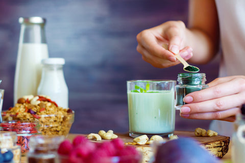 Person adding spirulina powder to smoothie with fruit, milk, and granola in background