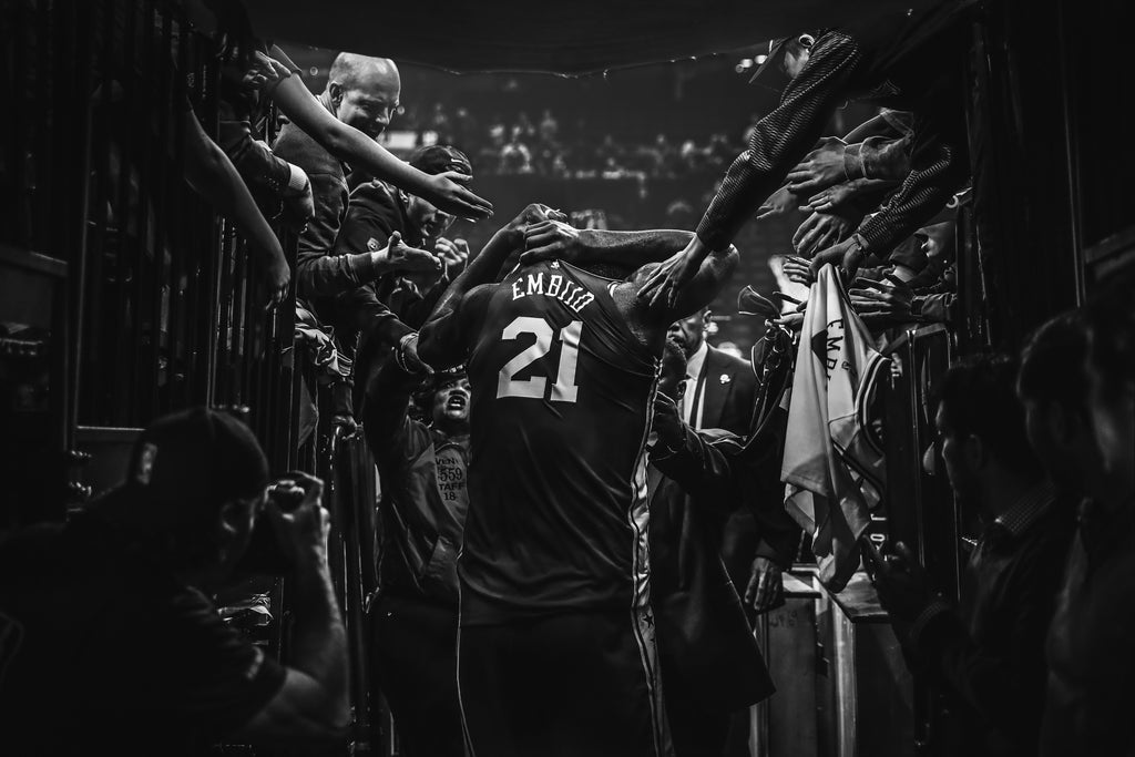 Philadelphia 76er Joel Embiid walks down the tunnel to the basketball court as fans reach over railing to give him high fives.