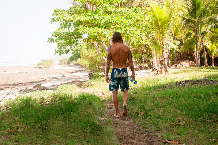 man walking through the jungle to the beach in costa rica, wearing riz swim shorts and carrying a glass water bottle