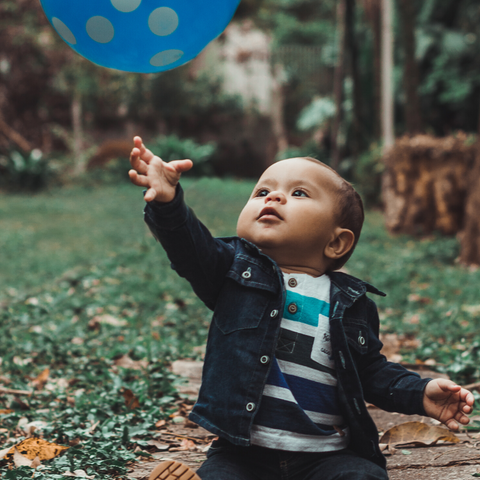 Baby boy wearing a denim jacket sits in a garden reaching up for a blue balloon