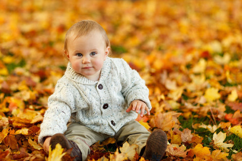 A baby boy wearing a cardigan sitting in a pile of autumn leaves