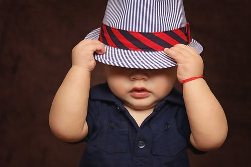 A baby boy wearing a navy top pulls a striped hat down over his eyes
