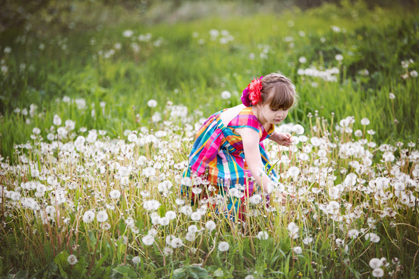 A young girl wearing a pink and blue dress is in a meadow of dandelion flowers