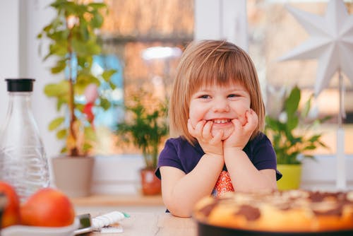 a toddler girl wearing a navy blue t-shirt is smiling sitting at a table surrounded by plants