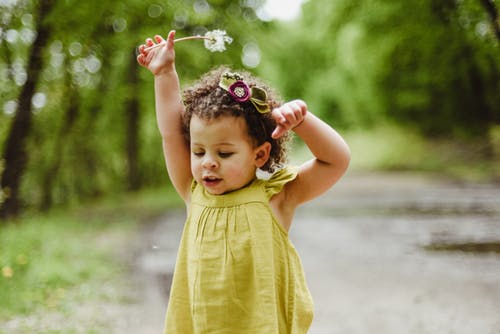 A toddler girl walking down a road wearing a yellow dress holds a dandelion above her head