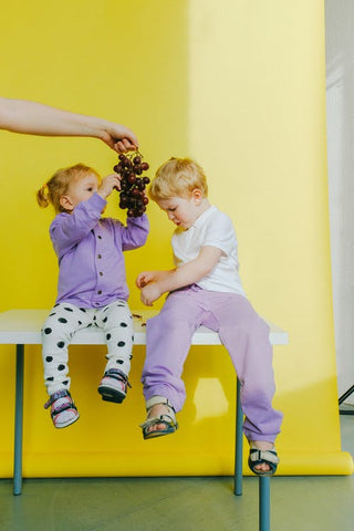 two young children wear matching colour unisex clothes holding a bunch of grapes