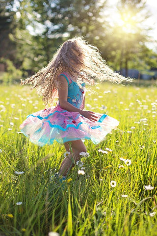 A young girl wearing a pink and blue dress with long curly hair spins around in tall grass on a sunny day