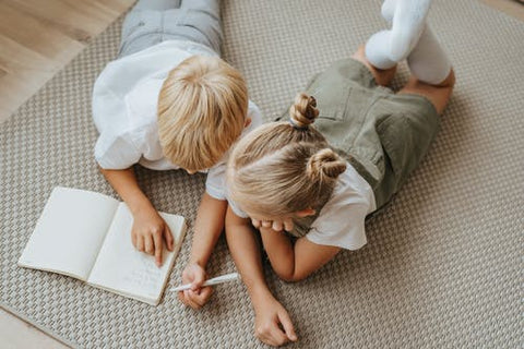 A boy and girl wear gender neutral clothing lay on the floor looking at a book