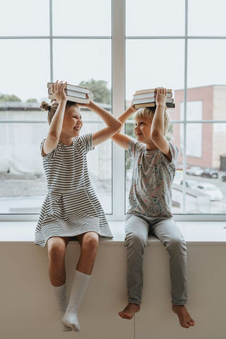 A boy and girl sit in the window with books on their heads