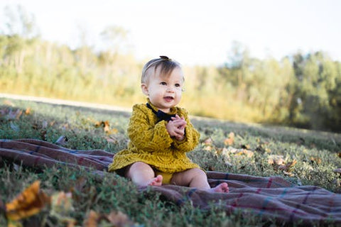 A baby girl in a yellow dress sits on a blanket on grass with leaves