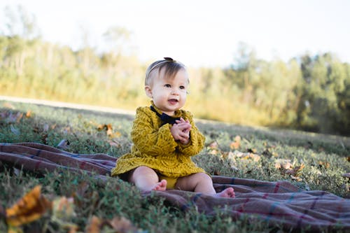 A baby girl wearing a yellow dress sits on a blanket laid on some grass