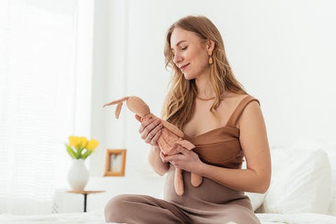 A pregnant lady dressed in brown sits on a white linen bed smiling holding a brown knitted soft toy