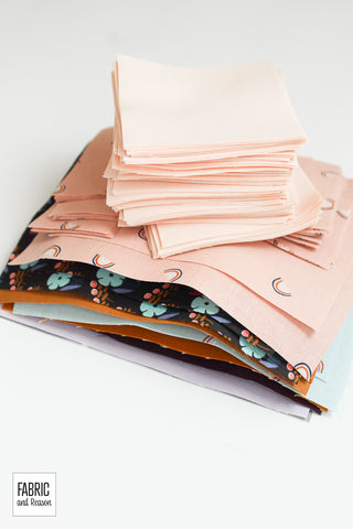 Stack of cut up fabric pieces in light pink, golden orange, light blue, and deep plum piled on a white surface.
