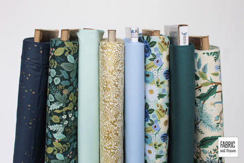 Stack of 8 bolts of fabric stood vertically on a white background in shades of deep teal, cream, gold metallic, and blue.