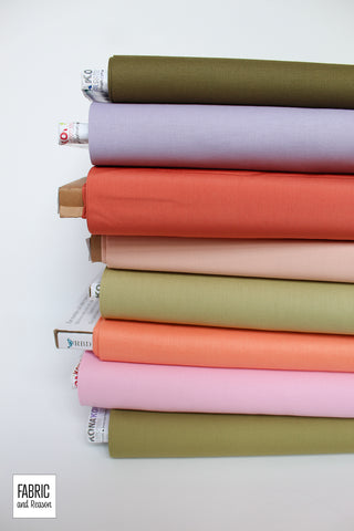 Eight bolts of fabric laid horizontally and stacked on a white background. Solid fabrics are in shades of green, orangey red, peach, pink, and lavender.