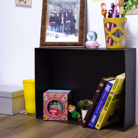 A living space with wooden floor and white wall showing a way to use giftopiia gift box as a storage space. A black giftopiia gift box is seen being used as a small box containing books, decorations, a toy, and more.