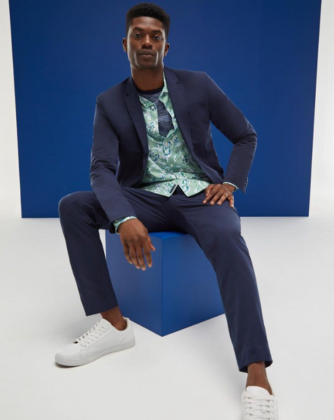 Man sitting on a blue cube wearing a blue suit, unbuttoned patterned dress shirt, heather blue tee, and white sneakers on a blue and white background