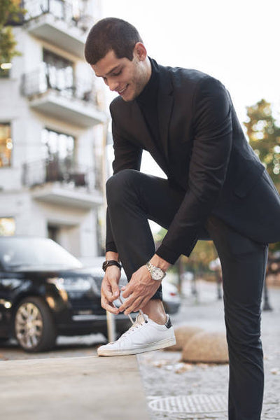 Man in a black suit, and turtleneck, tying his white sneakers. 