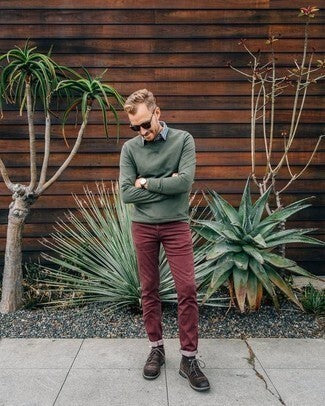  A man is standing in front of plants wearing red pants, a green sweater, brown boots, and sunglasses.