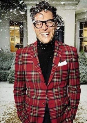 A man is standing in front of a snowy house laughing, wearing a black turtleneck, black pants, and a red plaid blazer.