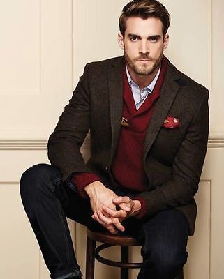 A man sitting on a stool wearing dark jeans, a white button up shirt and red sweater layered under a dark brown blazer.