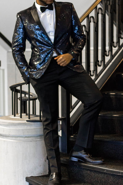 A man is standing on a black staircase wearing blazer pants, black sequin blazer, white dress shirt and a black bow tie.