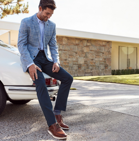 Man leaning against a white sports car wearing a blue plaid blazer with a blue button-up shirt, navy pants, and brown hybrid dress shoes