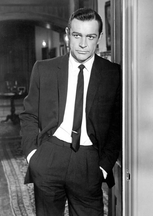 Sean Connery as james Bond leaning on a wall in a full black suit and slim black tie 