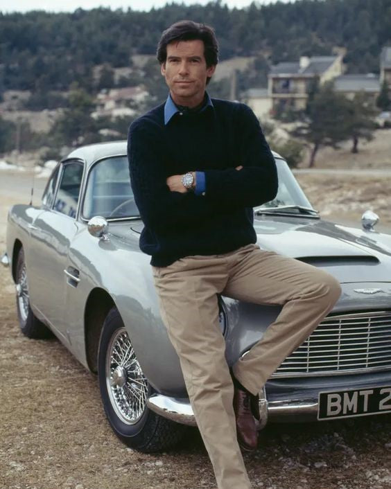 Pierce Brosman as james Bond sitting on the hood off a car in khaki chinos , a navy crewnwck sweater, and blue button-down