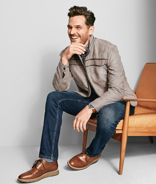 Man sitting in a chair sweater a taupe leather jacket, jeans, and tan hybrid dress shoes