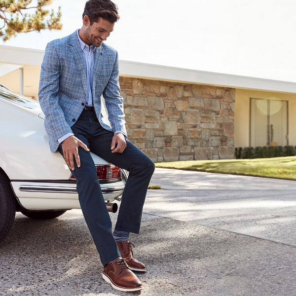 Man sitting on a car wearing a light blue button-down shirt, plaid blazer, jeans, and tan hybrid dress shoes