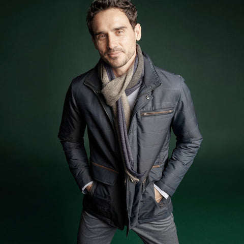 Man posing in a scarf, jacket, and pants against a solid green background.